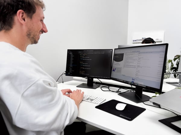 Ein Test Automation Engineer sitzt vor zwei Bildschirmen und einer Tastatur und lacht